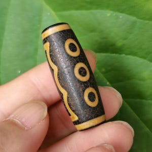 Op de afbeelding: Een cilindrische, donkerbruine dzi-kraal met drie cirkelvormige ontwerpen en een golvend patroon in een lichtere bruine kleur. De kraal wordt in de hand van een persoon gehouden tegen een groene bladeren achtergrond.