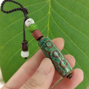 Puede incluir: Un colgante cilíndrico con un patrón jaspeado en verde y marrón con motivos circulares. Está unido a un cordón marrón oscuro con una cuenta blanca moteada, una cuenta verde sólida, una cuenta de madera roja oscura y una pequeña cuenta blanca.