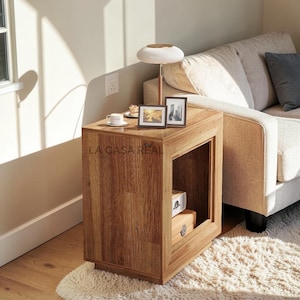May include: A wooden side table with a rectangular design and open storage. The table has a natural wood finish and features the text "LA CASA REAL". A small lamp, two picture frames, and a cup and saucer are on top.