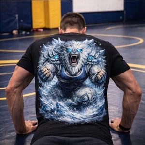 Könnte beinhalten: Schwarzes T-Shirt mit einem Grafikdesign eines brüllenden Yetis mit blauen Augen, umgeben von wirbelndem blauem Wasser und Eis. Das Wort "WRESTLING" ist im Hintergrund zu sehen, was auf ein Sportthema hindeutet.