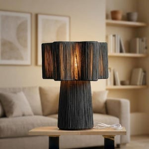 May include: A black table lamp with a textured shade and base, sitting on a wooden table. The lamp's shade has a layered design, and the base is a conical shape. A white power cord is visible.