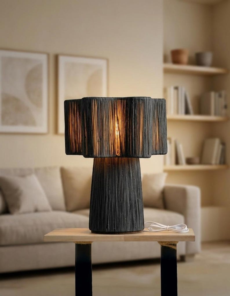 May include: A black table lamp with a textured shade and base, sitting on a wooden table. The lamp's shade has a layered design, and the base is a conical shape. A white power cord is visible.