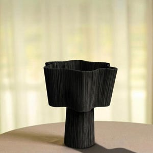May include: A black, textured vase with a unique, wavy rim. The vase has a pedestal base and sits on a neutral-colored surface. The design is modern and sculptural, suitable for home decor.