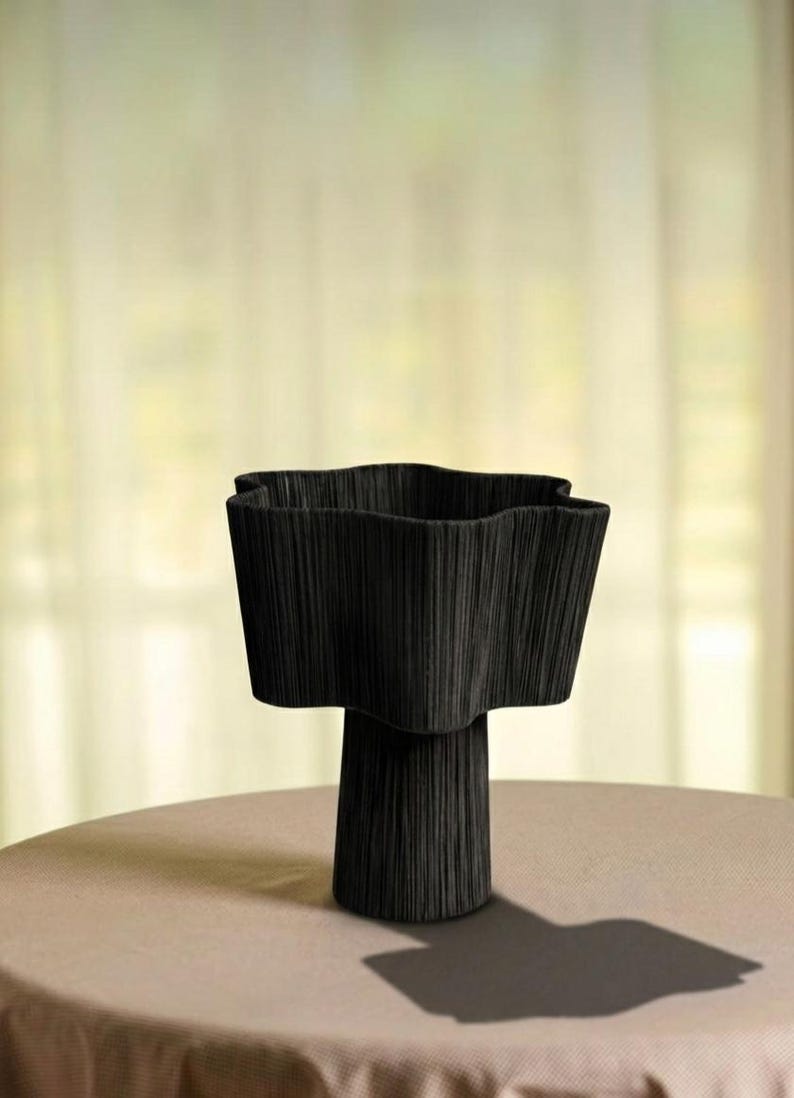 May include: A black, textured vase with a unique, wavy rim. The vase has a pedestal base and sits on a neutral-colored surface. The design is modern and sculptural, suitable for home decor.