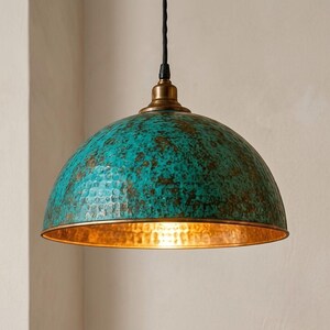 May include: A dome-shaped pendant light fixture featuring a hammered metal exterior with a weathered turquoise and bronze patina finish. The interior of the shade has a polished gold-toned metallic surface, suspended by a black fabric cord.