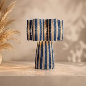 May include: A table lamp featuring a cylindrical base and a wide, scalloped shade, both wrapped in a textured rope material with alternating vertical stripes of deep blue and off-white, emitting a warm glow from the center of the shade.