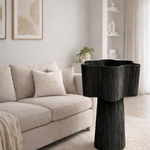 May include: A black, star-shaped accent table with a textured, vertical design. The table is set on a light-colored rug in a living room setting, with a beige sofa and framed artwork in the background.