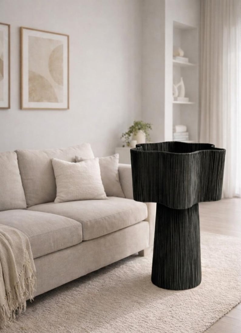 May include: A black, star-shaped accent table with a textured, vertical design. The table is set on a light-colored rug in a living room setting, with a beige sofa and framed artwork in the background.