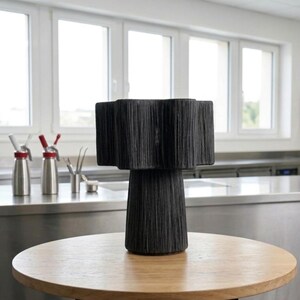 May include: A black decorative lamp with a textured, fringed shade and a cylindrical base. The lamp sits on a round, light-colored wooden table with a black metal base. The background includes windows and stainless steel surfaces.