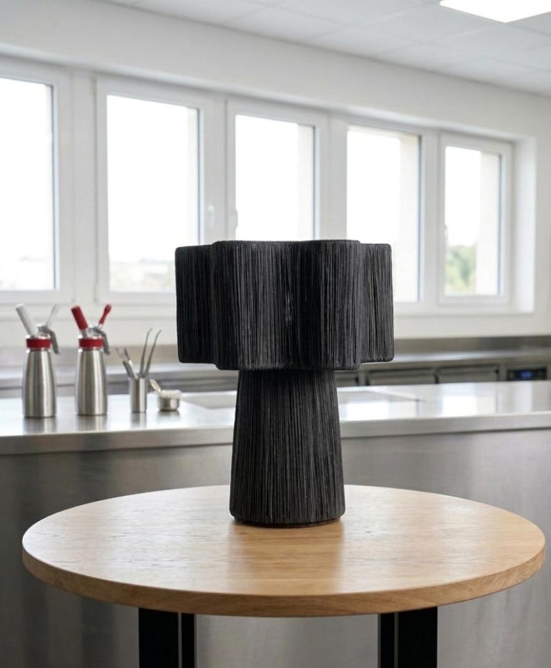 May include: A black decorative lamp with a textured, fringed shade and a cylindrical base. The lamp sits on a round, light-colored wooden table with a black metal base. The background includes windows and stainless steel surfaces.