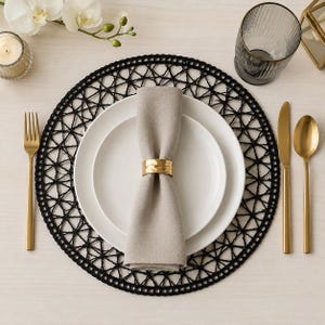 Puede incluir: Una mesa puesta con un mantel individual negro tejido, platos blancos y una servilleta beige sujeta por un anillo dorado. También hay cubiertos dorados y un vaso. Una vela y flores están en el fondo.