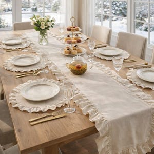 May include: A dining table set for a meal, featuring a light-colored wooden table with a cream-colored runner and placemats. White plates, gold cutlery, and clear glassware are arranged. A tiered dessert stand and a teapot are also present.