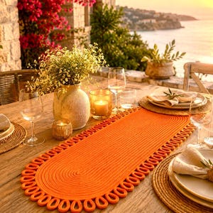 Puede incluir: Un camino de mesa ovalado de color naranja con textura trenzada y un borde decorativo de bucles. El camino está sobre una mesa de madera con platos blancos, servilletas beige, copas de vino, un jarrón con flores y dos velas pequeñas.