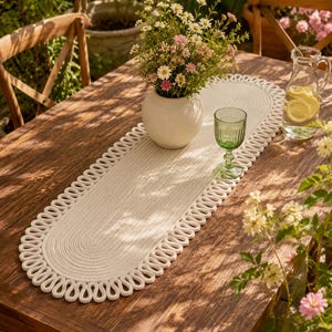 Pode incluir: Um caminho de mesa oval em tom branco sujo, feito de corda entrançada com uma borda decorativa em laços. O caminho está colocado sobre uma mesa de madeira e sustenta um vaso branco redondo com flores brancas e rosas, um copo verde e uma jarra.