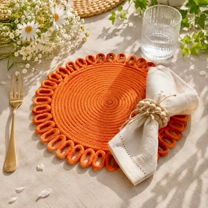 Può includere: Una tovaglietta rotonda intrecciata color arancione con bordo ad anelli, abbinata a un tovagliolo di lino color crema piegato e fissato con un portatovagliolo di perline di legno. Una forchetta in metallo dorato e un bicchiere rigato.
