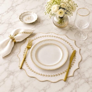 May include: A white scalloped placemat with gold leaf embroidery, a white plate with gold trim, a gold fork and knife, a white napkin with a gold leaf napkin ring, a small white dish, a glass vase with white flowers, and a ribbed glass.