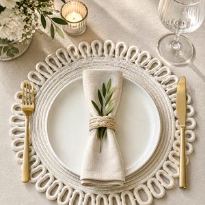 Op de afbeelding: Een rond gebroken wit geweven placemat met een lusvormige rand, voorzien van een wit keramieken bord, een beige linnen servet vastgehouden door een gevlochten touw servetring, een klein olijftakje, een gouden vork en een gouden mes.
