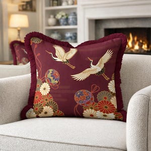 May include: Burgundy cushion with a Japanese-inspired design. It features flying cranes, floral patterns, and decorative balls. The cushion has a fringed edge and is on a light-coloured sofa.