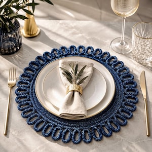 May include: A round, dark blue woven placemat with a looped edge design, featuring a white ceramic plate, a beige linen napkin, and a textured gold napkin ring. The setting includes a gold fork and knife, a glass, and a small decorative vase.