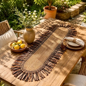 Op de afbeelding: Een ovale tafelloper gemaakt van gevlochten natuurlijke vezels, met een getextureerd beige midden en een decoratieve franjerand bestaande uit afwisselend donkerbruine en lichtbeige stroken, plat liggend op een houten tafeloppervlak.