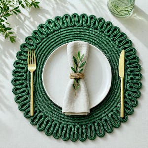 May include: A round, dark green woven placemat with a decorative looped edge, featuring a white ceramic plate, a beige linen napkin secured with a jute twine ring, a small green sprig, a gold-toned fork, and a gold-toned dinner knife.