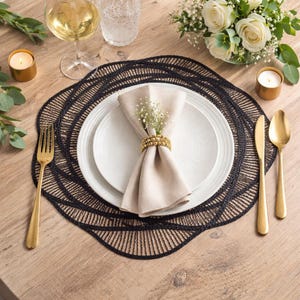 Könnte beinhalten: Tischarrangement mit einem schwarzen gewebten Tischset, weißen Tellern und goldenem Besteck. Eine beige Serviette wird von einem goldenen Perlen-Serviettenring gehalten. Ein Glas Wein und ein kleiner Strauß weißer Blumen sind ebenfalls vorhanden.