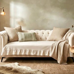 Puede incluir: Un sofá capitoné de color crema con brazos enrollados, que presenta una funda con faldón de volantes, dos cojines cuadrados grandes de color beige, un cojín rectangular a rayas verdes y blancas y una manta beige texturizada.