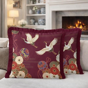 Puede incluir: Dos cojines burdeos con un diseño de inspiración japonesa. Cada cojín presenta un patrón repetido de grullas blancas en vuelo, elementos florales y bolas decorativas. Los cojines tienen un borde con volantes.