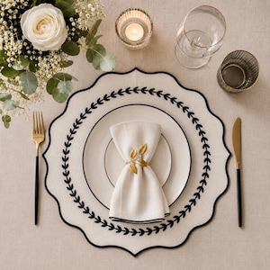 May include: Elegant table setting with a white plate, napkin, and gold leaf napkin ring. A black and gold fork and knife are on either side of the plate. A white scalloped placemat with black trim and a floral arrangement complete the setting.