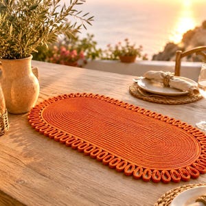 Puede incluir: Un mantel individual ovalado de color naranja vibrante con un centro enrollado apretado y un borde decorativo de bucles. El mantel está construido con un material de cordón grueso y texturizado, creando un patrón distintivo sobre la mesa.