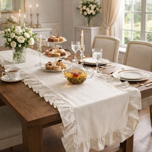 May include: A dining table set for tea with a cream-colored ruffled table runner. The table is set with white plates, glasses, and silverware. A glass teapot with a blooming flower sits on the runner, along with a tiered dessert stand.