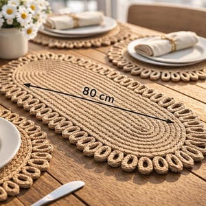 Puede incluir: Un camino de mesa ovalado tejido, de 80 cm de largo, con un borde festoneado, exhibido sobre una mesa de madera. También se ven manteles individuales y platos a juego con servilletas, junto con un jarrón de flores blancas y saleros y pimenteros. La escena sugiere un comedor.