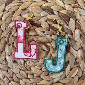 May include: Two fabric letter keychains featuring gold-tone lobster clasps. The letter L is white with a red strawberry pattern and pink stitching. The letter J is light blue with a white daisy pattern and dark teal stitching.