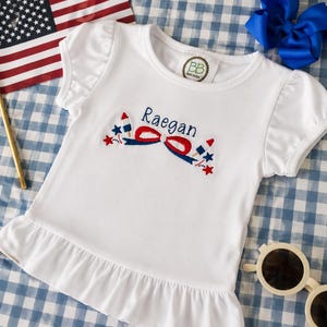 Puede incluir: Una camiseta blanca de manga corta con dobladillo con volantes y mangas abullonadas. El pecho presenta un diseño bordado con el nombre Raegan en azul, acompañado de un lazo rojo y azul, estrellas pequeñas y dos polos en forma de cohete.