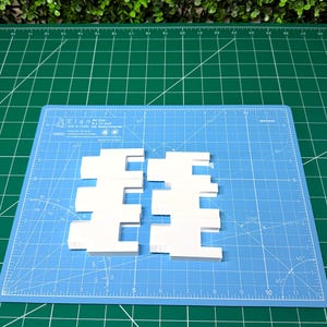 Könnte beinhalten: Zwei identische weiße Schaumstoffteile mit einem gekerbten, ineinandergreifenden geometrischen Design. Jedes Teil weist auf einer Seite drei rechteckige Vorsprünge und auf der anderen Seite entsprechende Vertiefungen auf, liegend auf einer blauen Schneidematte.