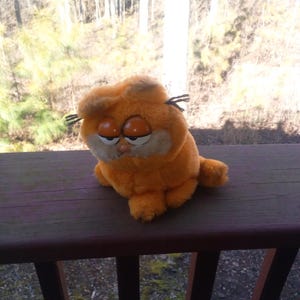 May include: An orange plush Garfield toy with a grumpy expression. The stuffed animal has black whiskers and is sitting on a wooden railing. The background shows a view of trees and foliage.