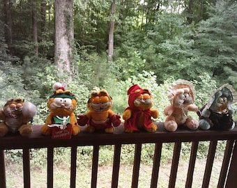 May include: A collection of six Garfield plush toys arranged in a row. The figures include a cat with a shopping bag labeled SHOP TIL YOU DROP, a cat in a red cape, a cat in a bonnet, and a cat with a pacifier, all featuring orange fur.