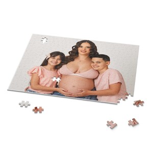 May include: A jigsaw puzzle featuring a family portrait of a pregnant woman and two children. The woman wears a pink top, and the children wear pink shirts. The puzzle is mostly white with a few puzzle pieces scattered around.