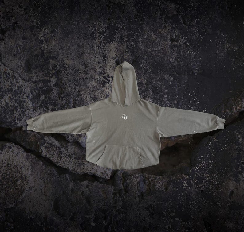 May include: A sage green hooded sweatshirt featuring long sleeves, a front kangaroo pocket, and a small white graphic logo centred on the chest. The garment has a relaxed fit with ribbed cuffs and a matching hood, showcasing a subtle distressed finish.
