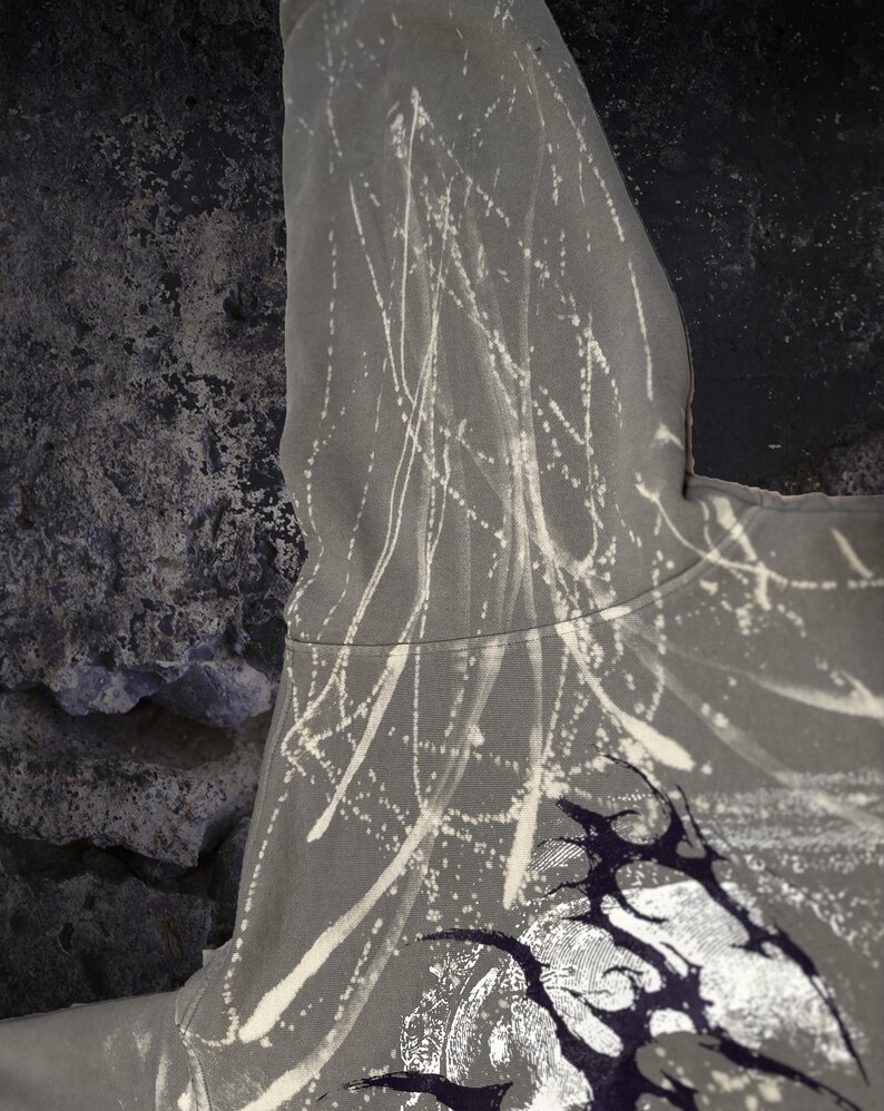 May include: A grey hoodie featuring a bleached, abstract splatter pattern of white lines and streaks across the fabric. The garment includes a dark, intricate graphic design near the bottom edge, showcasing sharp, pointed shapes in black and white.