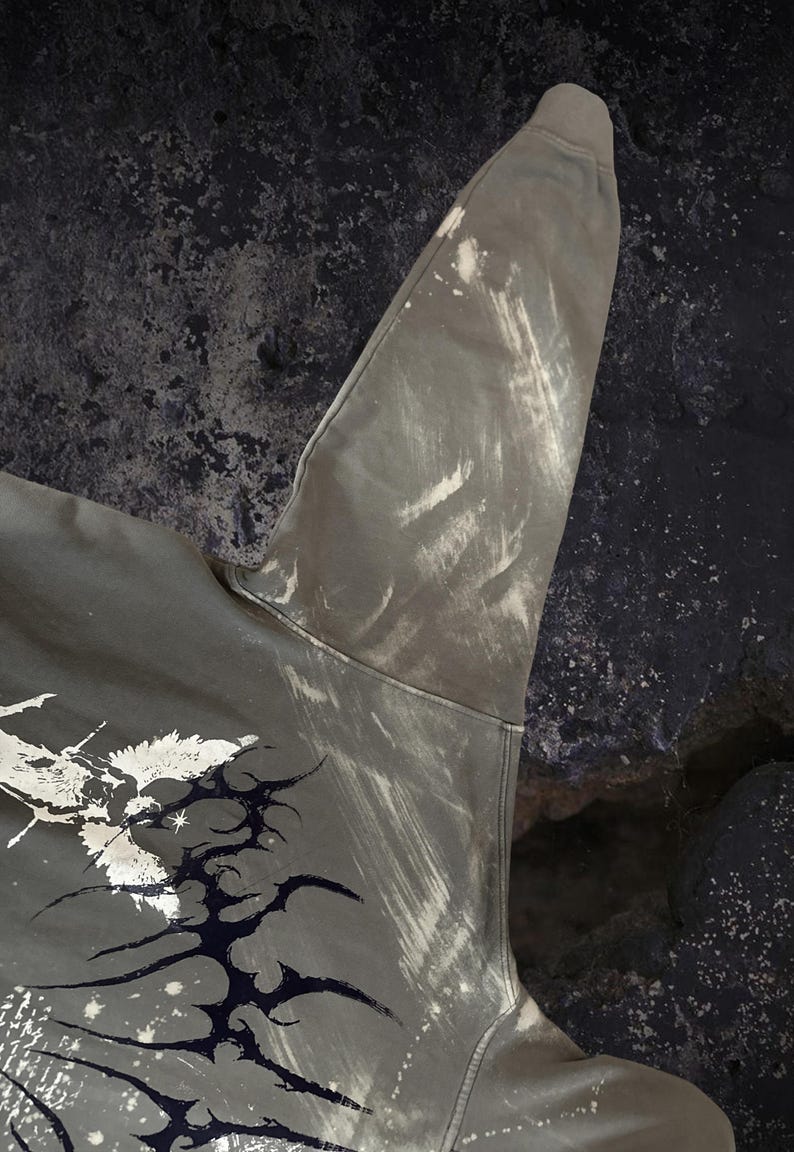May include: An olive green long-sleeve sweatshirt featuring a distressed, bleached effect on the sleeve and shoulder. The garment displays a dark, intricate, abstract graphic print with sharp, thorn-like lines and white splattered accents.