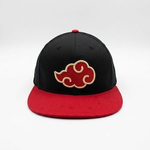 Könnte beinhalten: Eine schwarze Baseballkappe mit flachem rotem Schirm. Auf der Vorderseite befindet sich eine gestickte rote Wolkenform mit einer dicken beigen Umrandung. Die Kappe hat einen kleinen roten Knopf oben und schwarze Ösen zur Belüftung an den Seiten.