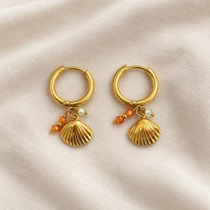 Peut inclure: Une paire de créoles dorées avec des breloques pendantes. Chaque boucle d'oreille comporte un pendentif en forme de coquillage métallique accompagné de petites perles orange à facettes et d'une perle vert pâle fixée à l'anneau.