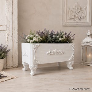 Peut inclure: Un bac à fleurs rectangulaire blanc avec des détails ornés et des pieds incurvés, rempli de verdure et de fleurs. La boîte présente des moulures décoratives et est placée près d'une lanterne blanche et d'une plante en pot.