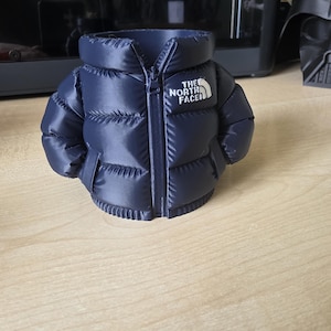 May include: A navy blue miniature puffer jacket with a zipper and the text "THE NORTH FACE" in white. The jacket is a decorative item, possibly a container or holder, and sits on a wooden surface.
