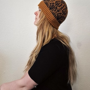 Op de afbeelding: Een gebreide muts in bruin- en zwarttinten met een geometrisch patroon. De muts heeft een geribbelde rand. De persoon op de foto draagt een zwarte top en heeft lang blond haar.