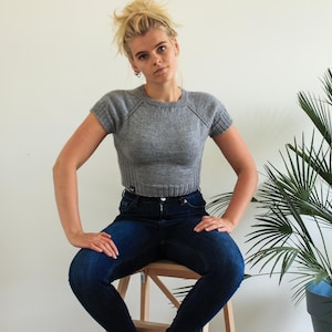 May include: A gray, short-sleeved crop top and dark blue jeans. The top is a knit fabric. The person is sitting on a wooden stool. A green plant in a blue pot is to the right.