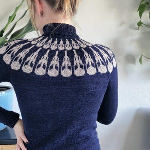 Op de afbeelding: Marineblauwe coltrui met een beige geometrisch patroon over de schouders. De trui is nauwsluitend gebreid met lange mouwen. Het haar van het model is in een knot.
