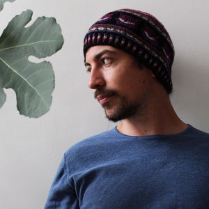 Op de afbeelding: Een gebreide beanie met een patroon in tinten bordeauxrood, marineblauw en wit. De hoed wordt gedragen door een persoon die een blauw shirt met lange mouwen draagt. Een vijgenblad is zichtbaar op de achtergrond.