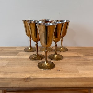 Puede incluir: Seis copas doradas vintage con acabado metálico dispuestas sobre una mesa de madera. Las copas tienen un diseño clásico con un cuenco acampanado, un tallo delgado y una base redonda. El fondo es una pared lisa de color blanquecino.